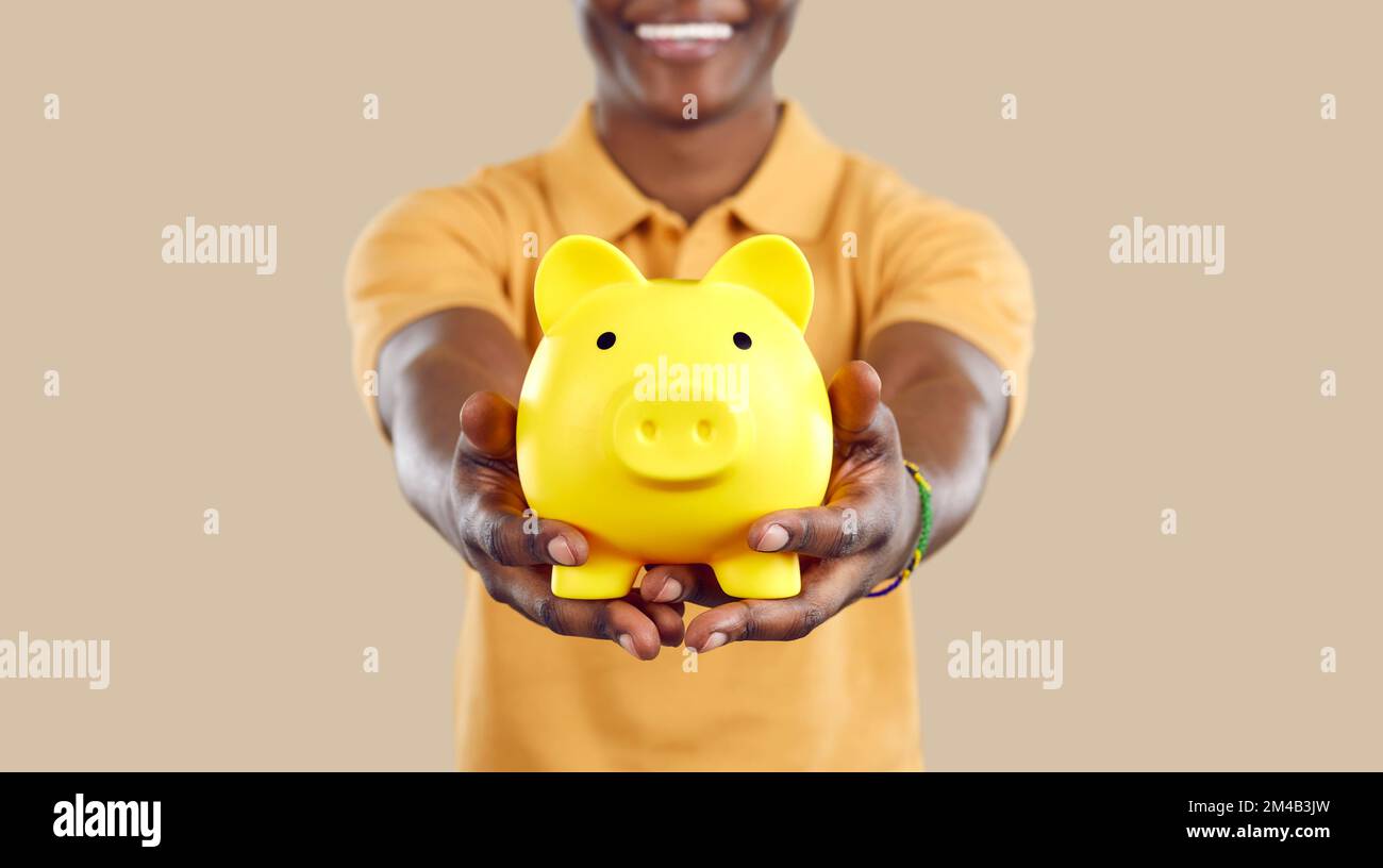 Schweinebank in Form eines gelben Schweines in den Händen eines lächelnden schwarzen Mannes auf beigem Hintergrund. Stockfoto