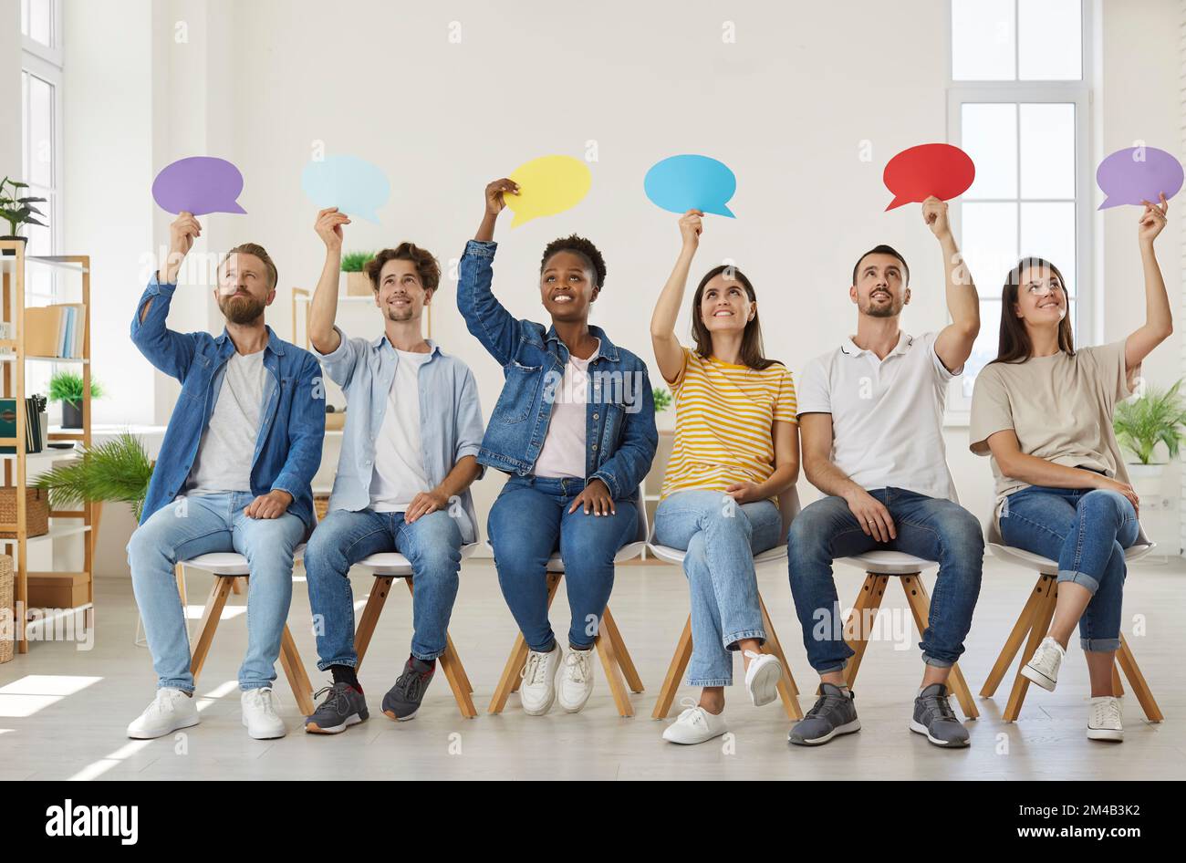Positive, vielfältige Menschen sitzen in der Reihe und äußern ihre Meinung, indem sie farbige Sprechblasen erzeugen. Stockfoto