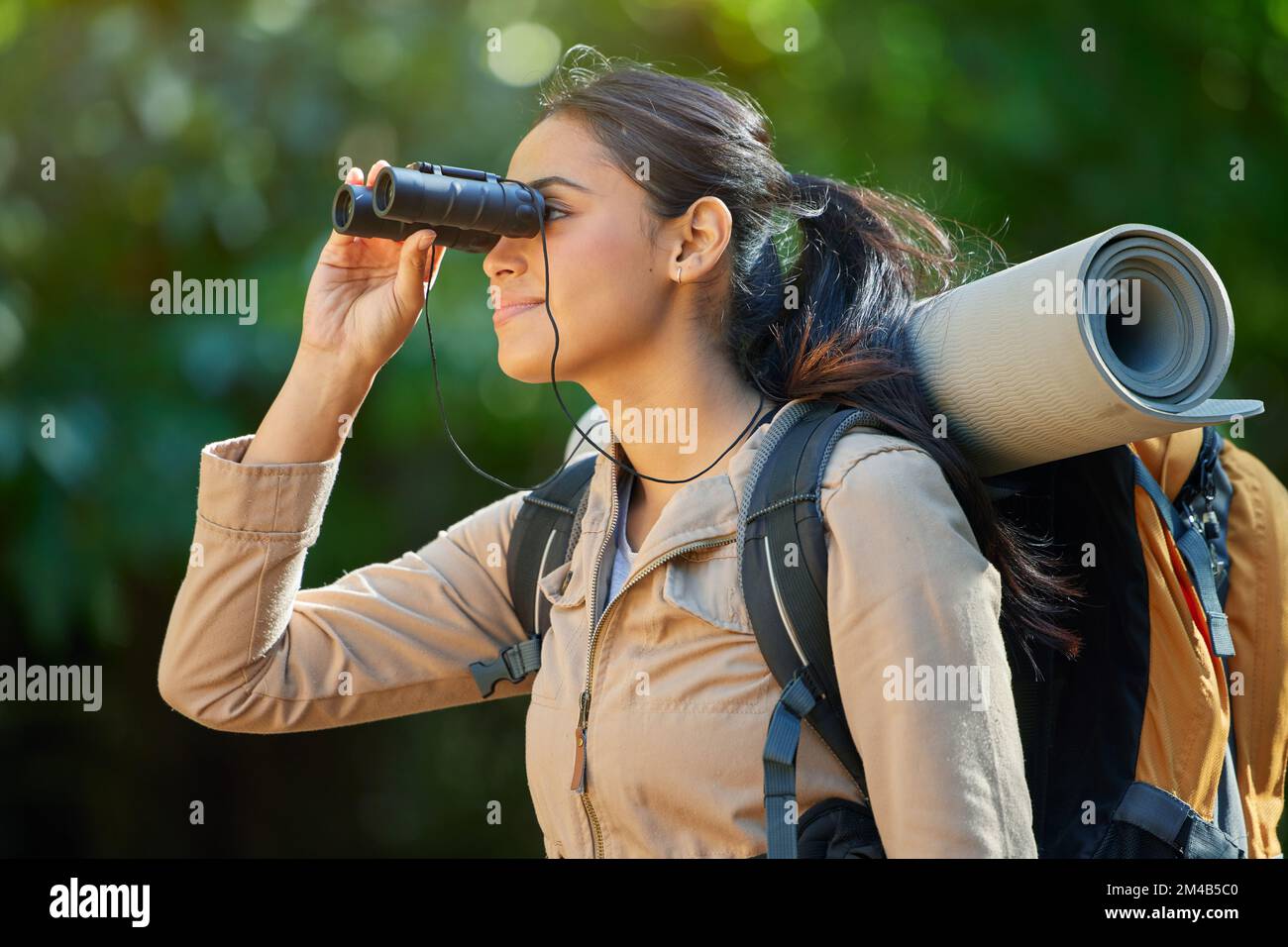 Wandern, Fernglas und Sehvermögen im Wald mit Rucksack, Campingausrüstung und Lächeln auf der Naturreise. Glückliches Wanderer-Mädchen, Vogelbeobachtung und Trekking Stockfoto
