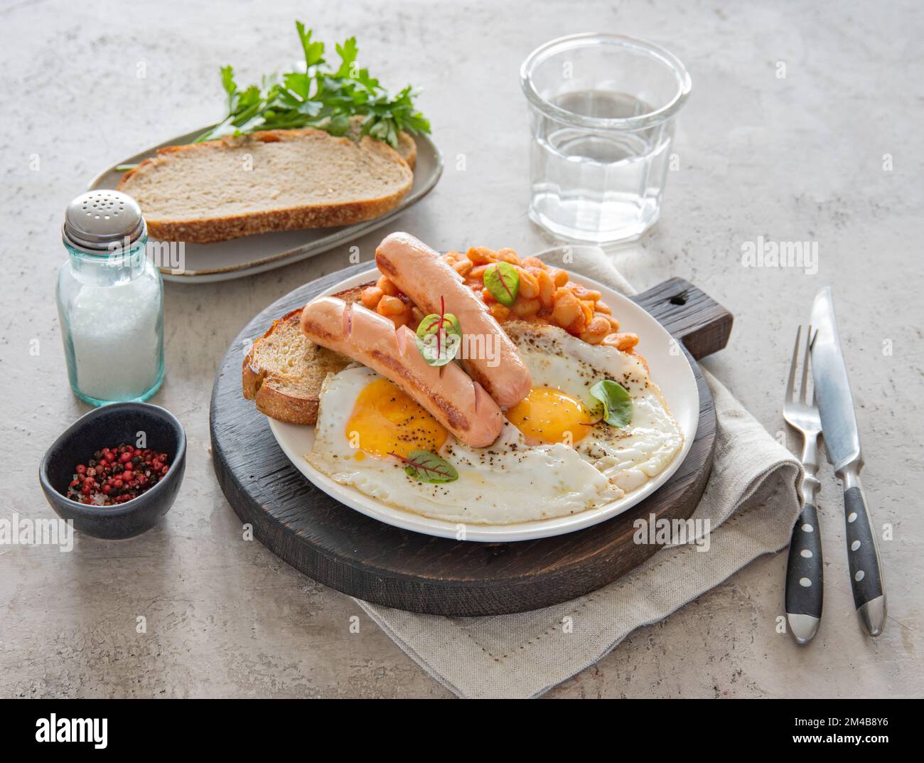 Traditionelles englisches Frühstück mit Spiegeleiern, Würstchen, Bohnen, frischen Kräutern, rotem Pfeffer und Toast auf grauem Hintergrund. Konzept für Morgenessen. Stockfoto