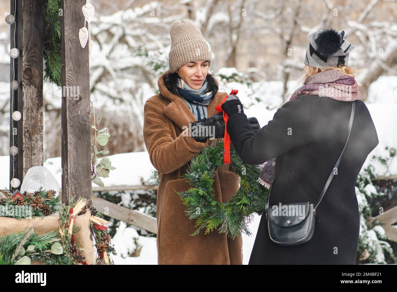 Wunderschöne Mädchen in warmer Kleidung, die im Winter auf einem Straßenmarkt wunderschöne Weihnachtsgirlande oder Kranz kaufen, mit Schnee und Rauch von warmen Speisen Stockfoto