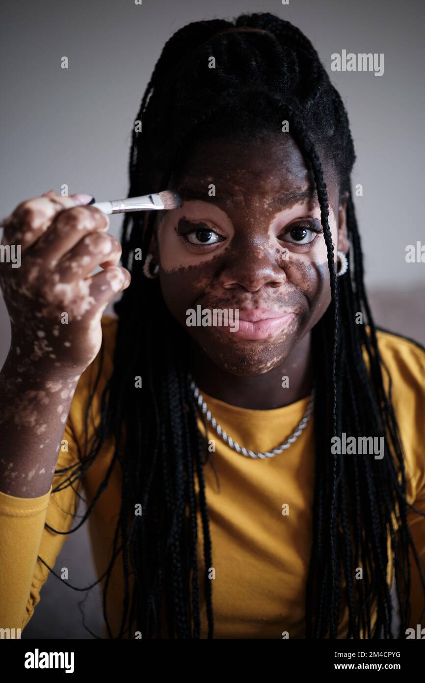 Lächelnde schwarze Frau mit Vitiligo, die sich mit Pinsel schminkt Stockfoto
