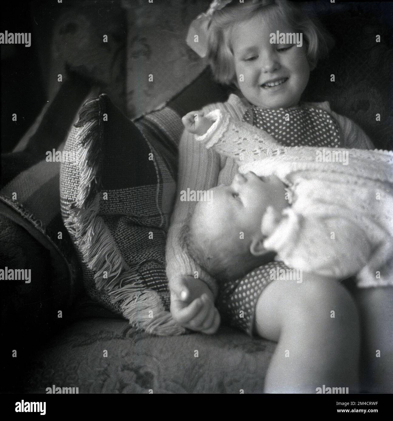 1960er Jahre, historisch, ein junges Mädchen, das mit ihrer Schwester auf einem Sofa sitzt, in einem Baumwolloutfit, auf ihrem Schoß liegend. Stockfoto