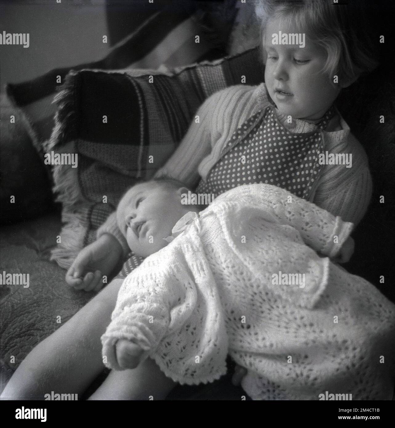 1960er Jahre, historisch, ein junges Mädchen, das mit ihrer Schwester auf einem Sofa sitzt, in einem Baumwolloutfit, auf ihrem Schoß liegend. Stockfoto