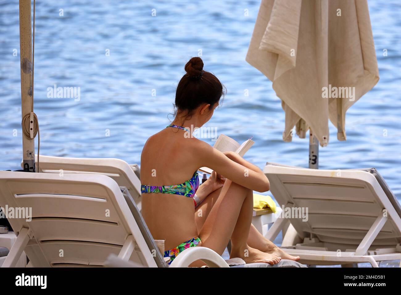 Ein Mädchen im Bikini liest Papierbücher und sitzt auf einem Liegestuhl am Strand. Entspannen Sie sich im Luxusresort Stockfoto