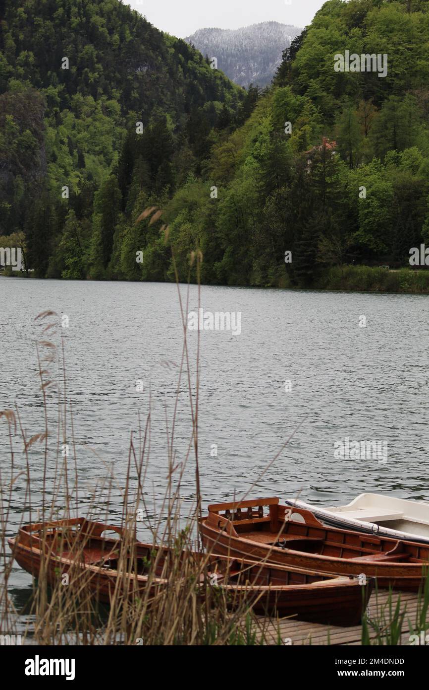 An einem regnerischen Tag im Hintergrund liegt ein Holzboot auf einem See mit verschneitem und grünem Wald. Stockfoto