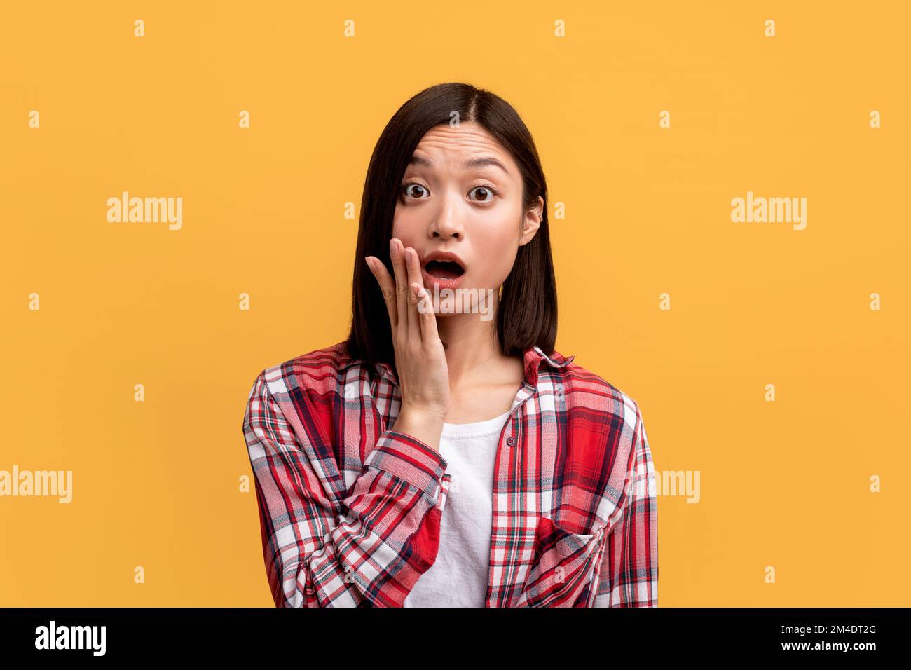 Schockierte junge Asiatin, die mit offenem Mund in die Kamera schaut und die Wange berührt, emotional auf Nachrichten reagiert Stockfoto