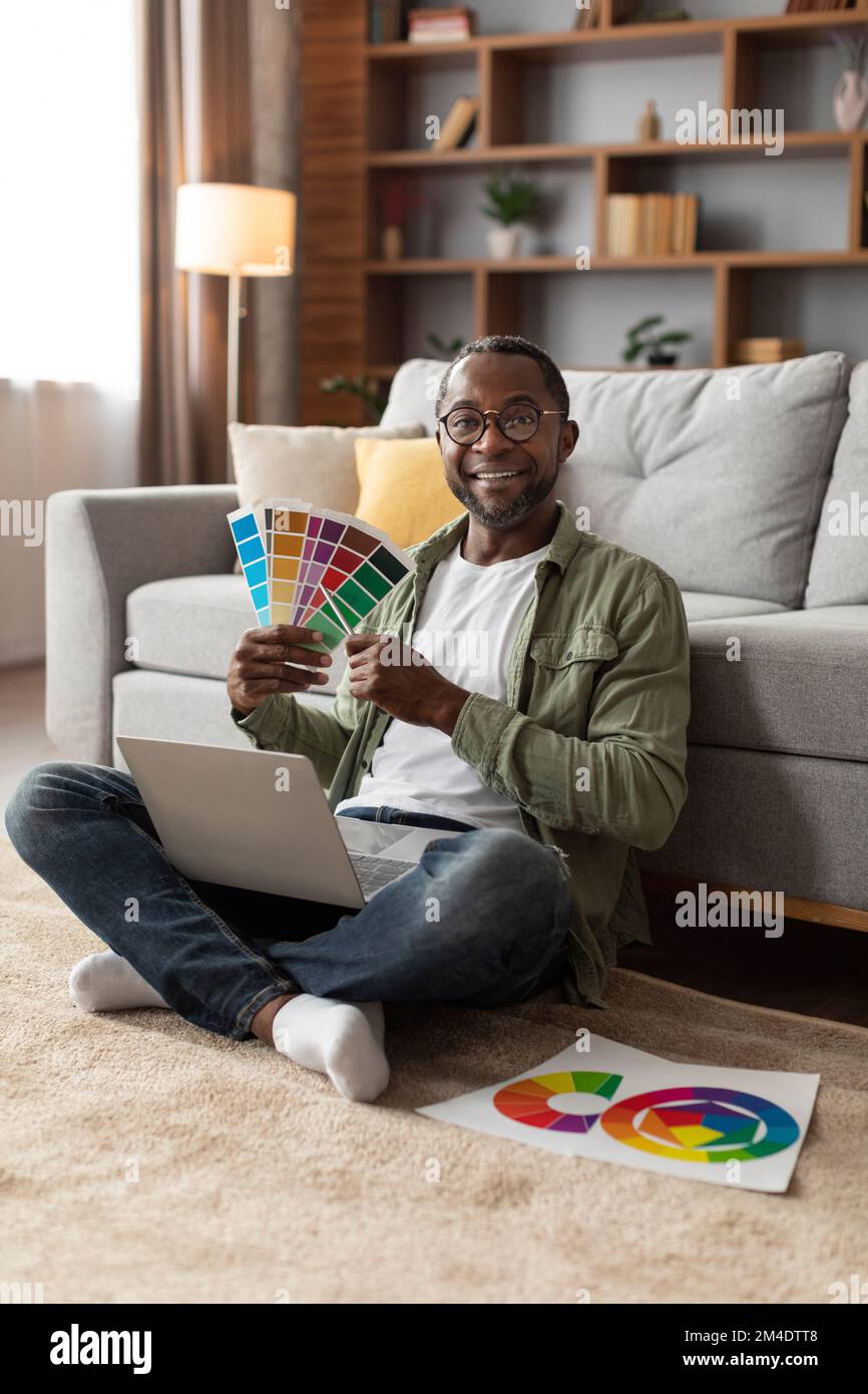 Der fröhliche Erwachsene Schwarze mit Brille benutzt einen Computer, arbeitet mit Farbpaletten für Design, sitzt auf dem Boden Stockfoto