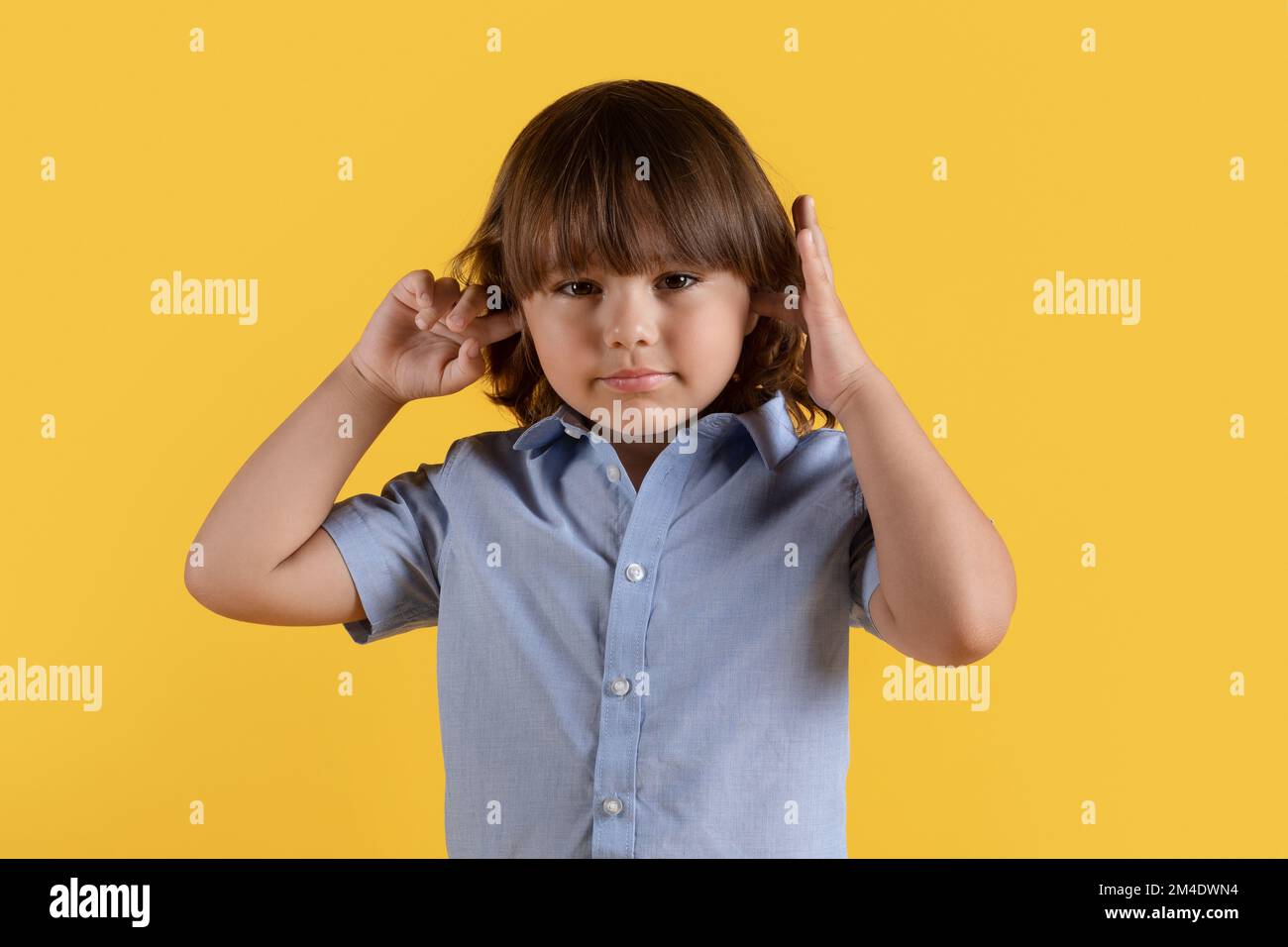 Höre nichts Böses. Stur, kleiner Junge, der die Ohren mit Händen schließt, nicht zuhören will, in die Kamera schaut, orangefarbener Hintergrund Stockfoto