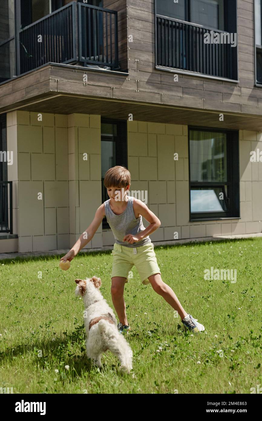 Ein langes Porträt eines lächelnden Jungen, der mit einem süßen Hund auf dem Rasen im Sonnenlicht spielt Stockfoto