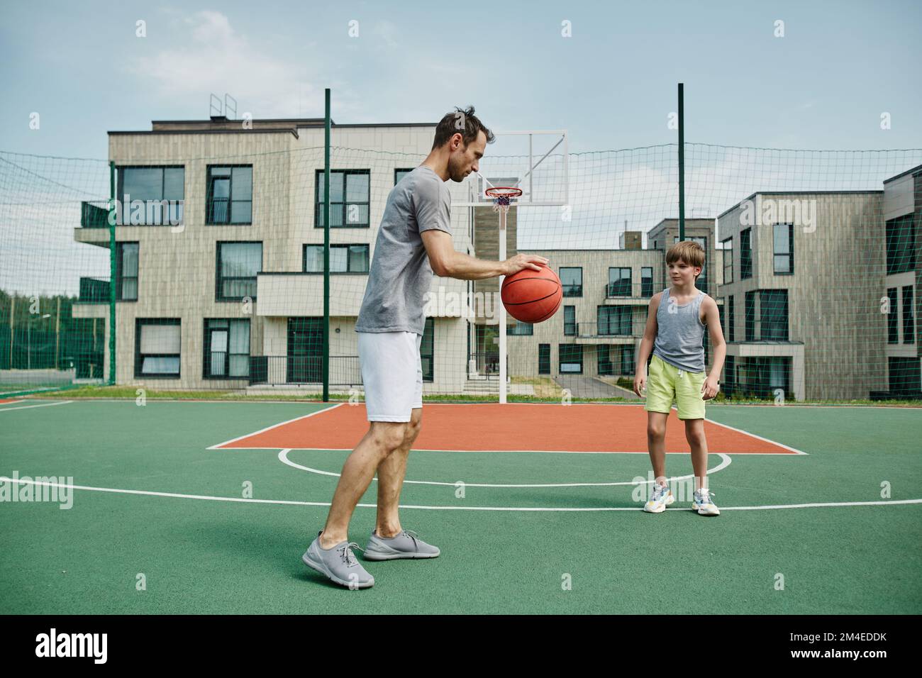 Ganzkörperporträt von Vater und Sohn, die gemeinsam im Freien Basketball spielen, in einem modernen Wohnkomplex Stockfoto