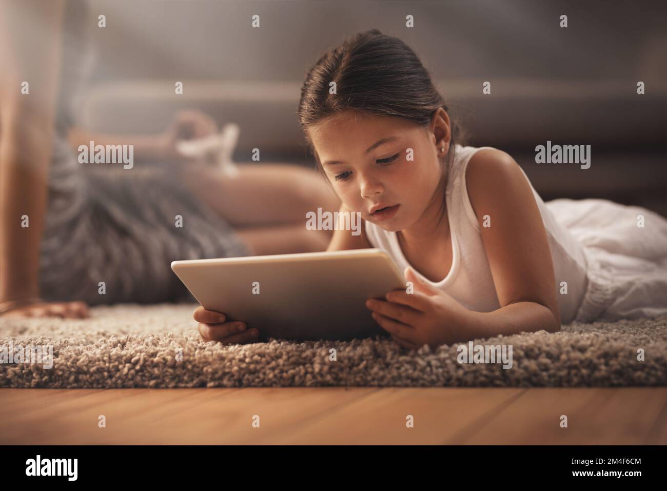 Eine App pro Tag hält Langeweile fern. Ein bezauberndes kleines Mädchen, das zu Hause ein digitales Tablet auf dem Boden mit seiner Mutter im Hintergrund benutzt. Stockfoto