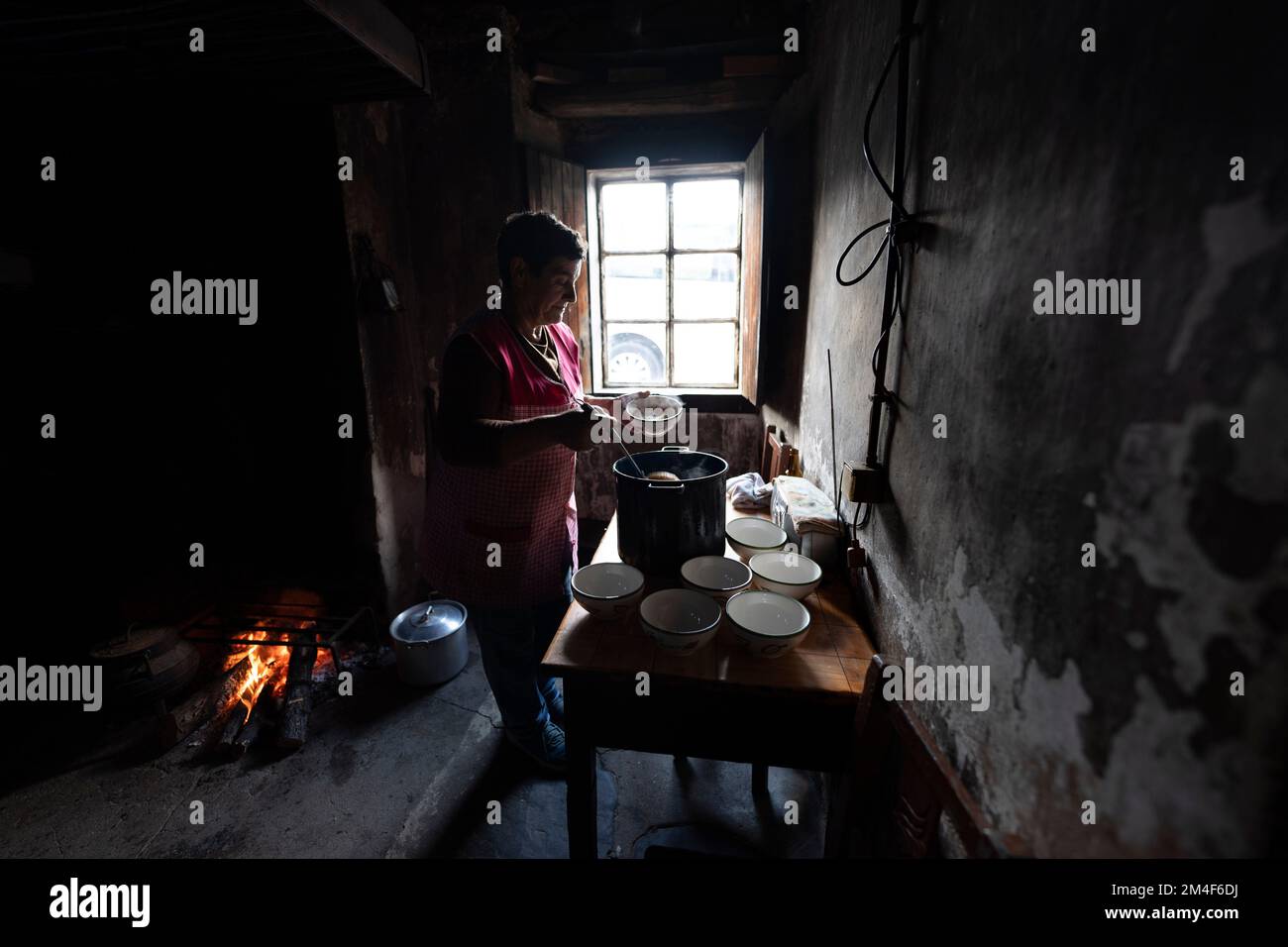 Alte Frau, die am offenen Feuer in einer kleinen, schwach beleuchteten Küche in einem Landhaus kocht Stockfoto
