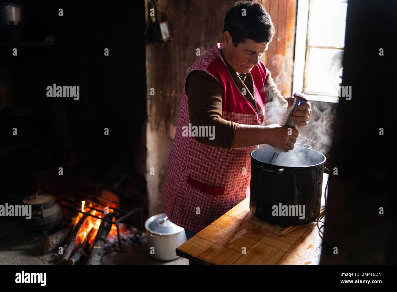 Alte Frau, die am offenen Feuer in einer kleinen, schwach beleuchteten Küche in einem Landhaus kocht Stockfoto