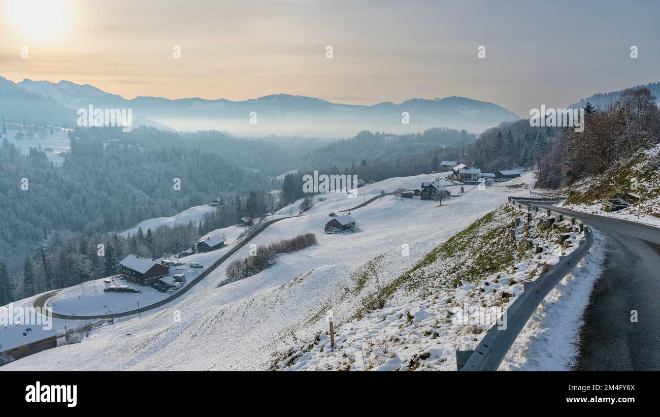 Frisch verschneite Bergstraße, die sich ins Tal schlängelt, Bauernhaus am Straßenrand, Berge und Täler mit Wäldern und Wiesen im Hintergrund, Abend Stockfoto