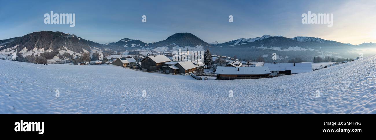 Winterliches Panorama mit verschneiter Landschaft, Blick über Dorf im Winter mit Schnee auf Wiese und Wälder, Berge des Bregenzerwald im Hintergrund, Stockfoto