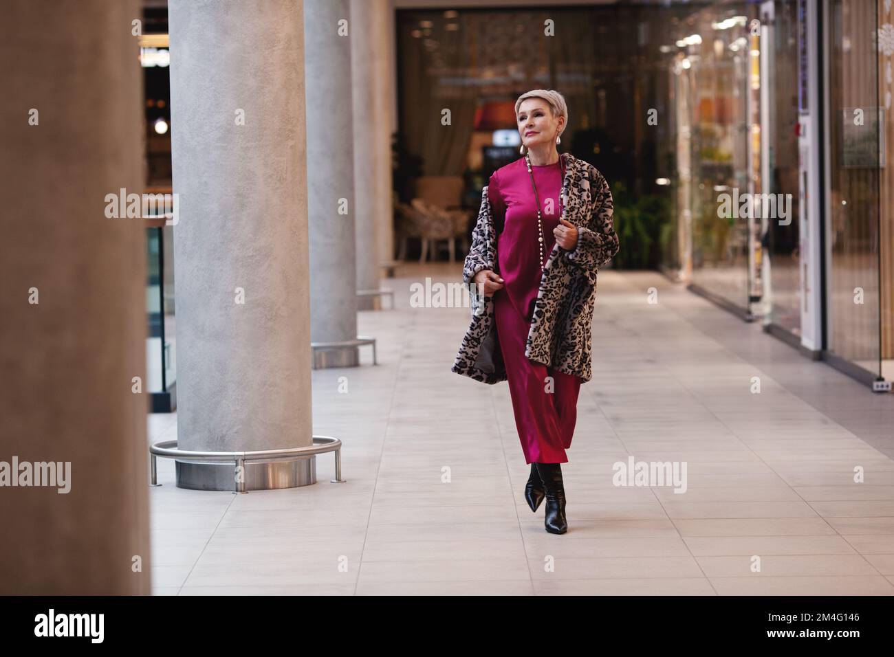 Wunderschöne Frau mittleren Alters, die modische Kleidung trägt langes burgunderfarbenes Seidenkleid, Mantel mit Leopardenmuster, schwarze Stiefel. Elegante Model Walking-Frau Stockfoto
