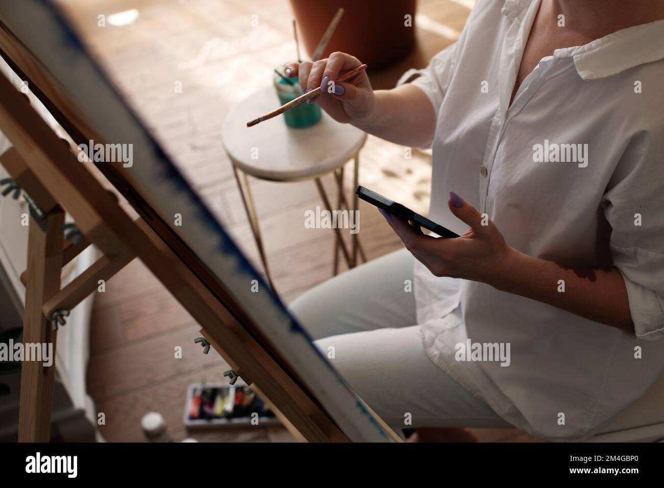 Eine Künstlerin zeichnet Bilder mit einem Pinsel auf der Staffelei und hält ihr Smartphone in der Hand. Sie sieht sich Online-Kunstunterricht an Stockfoto