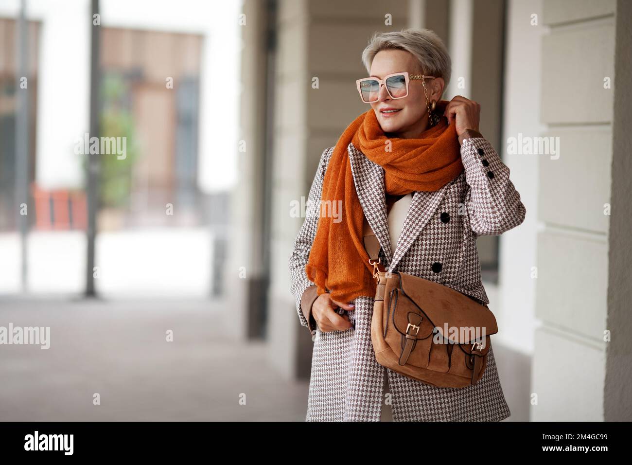Lächelnde Frau Im Außenporträt. Das kurzblonde Modemodell trägt stilvolle Kleidung, eine zweireihige Jacke, eine Lederhandtasche, einen ockerfarbenen Strickschal und Stockfoto