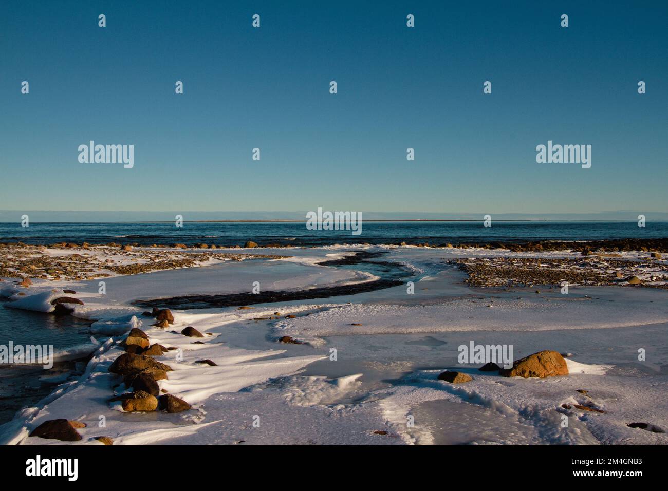 Arktische Landschaft - ein kleiner Bach, der in ein größeres Gewässer fließt, umgeben von gefrorener arktischer Tundra mit schneebedeckten Felsen an einem klaren kalten Tag in der Nähe Stockfoto