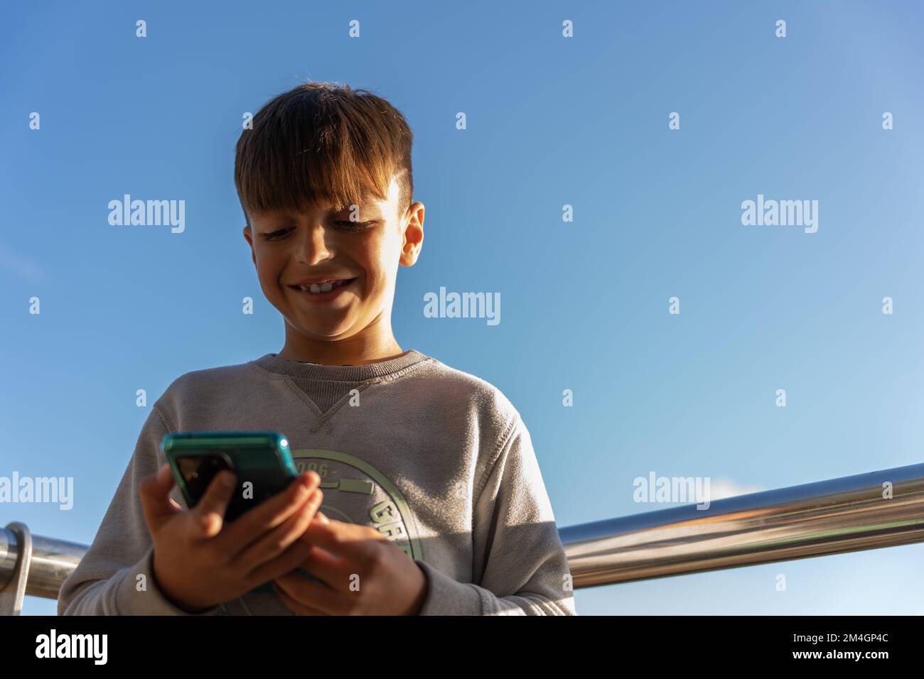 Von unten fröhlicher Junge in legerer Kleidung, der lächelt und Videos auf dem Handy ansieht, während er Zeit auf der Terrasse vor dem wolkenlosen blauen Himmel verbringt Stockfoto