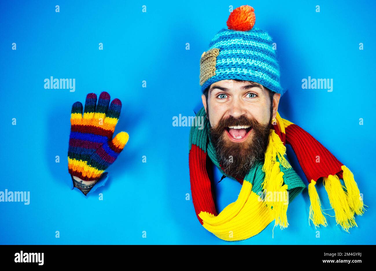 Bärtiger Mann mit farbenfroher Mütze, Schal und Handschuhen, die Hand winken. Winterkleidung. Strickmode für Männer. Stockfoto
