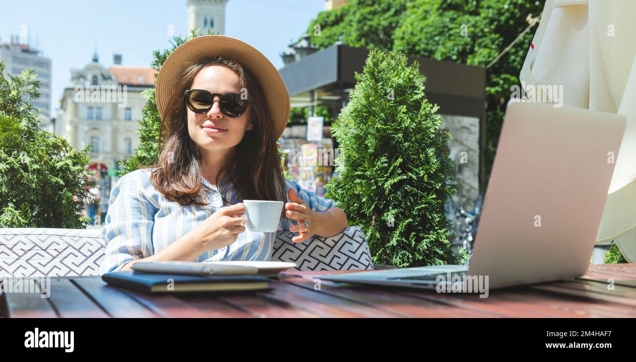 Junge Erwachsene Frau mit Sonnenbrille und Strohhut, die im Café im Freien sitzt und während der Geschäftsreise Kaffee trinkt. Stockfoto