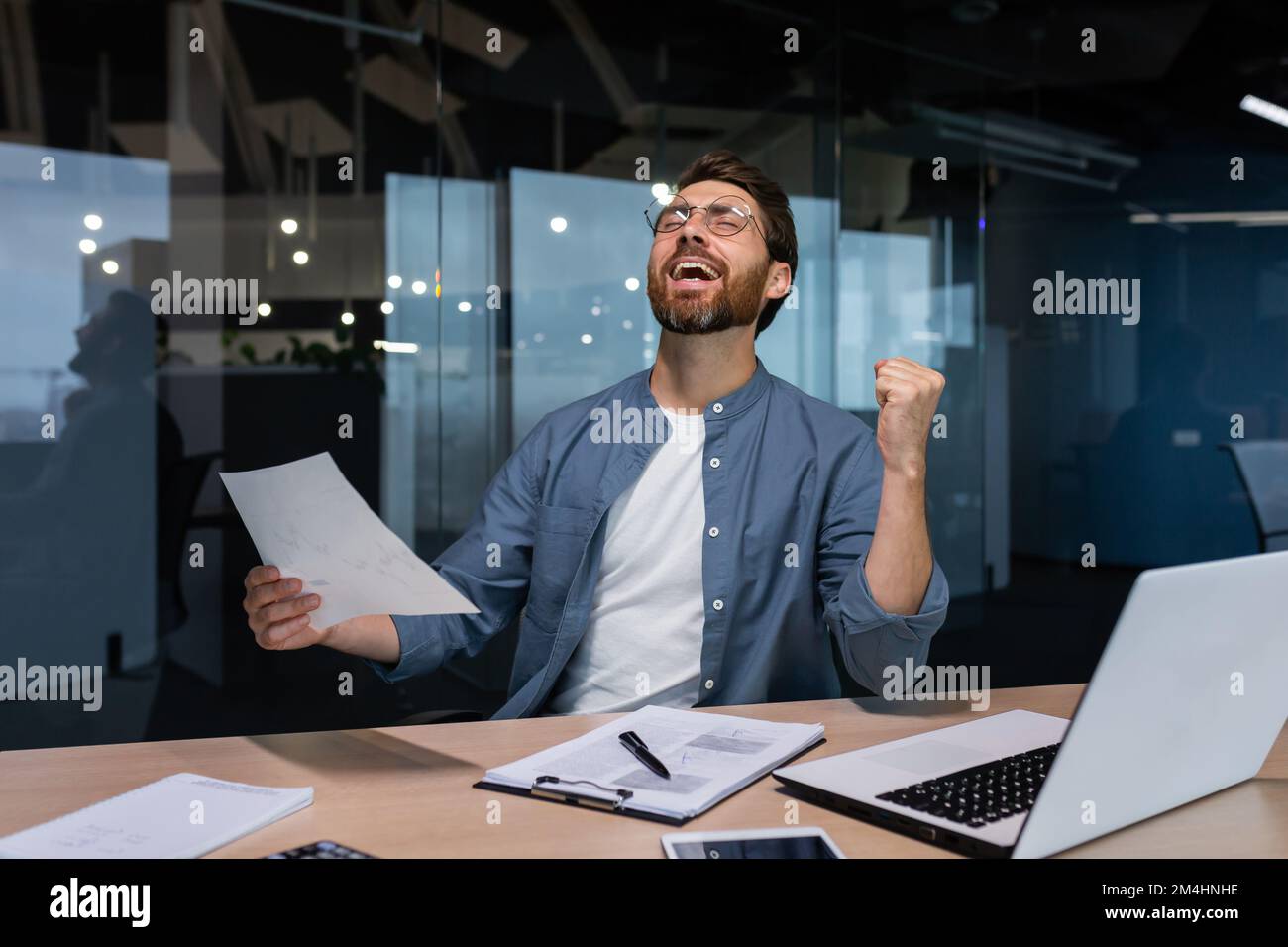 Erfolgreicher Geschäftsmann in legerem Hemd, der Papierkram erledigt, Chef mit Bart und Brille, der am Arbeitsplatz mit einem Laptop am Schreibtisch sitzt und Dokumente bearbeitet, die Hand hochhält und Sieg und Triumph feiert. Stockfoto