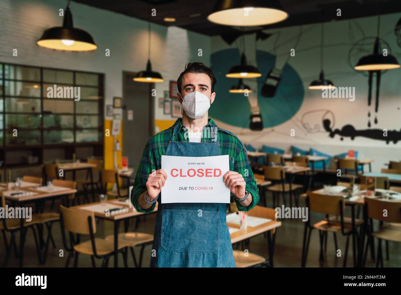 Ein Mann mit einer schützenden Gesichtsmaske hält ein Brett mit "Sorry, dass wir geschlossen sind" -Schild in einem Café. Konzept der Schließung von Unternehmen durch Covid-19-Virus und qua Stockfoto