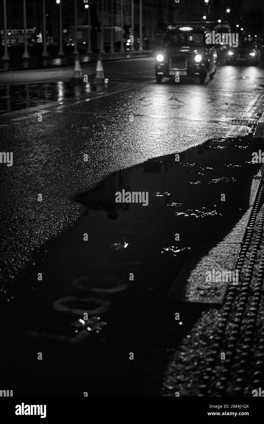Schwarzweiß-Schwarzweißbild mit verschwommenem Hintergrund eines schwarzen Londoner Fahrerhauses auf nasser Straße mit großer Pfütze Stockfoto