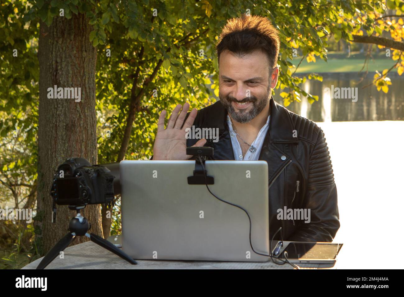 Ein Junge im Livestreaming, der seine Follower über die Webcam seines Laptops begrüßt, der im Freien zwischen den Bäumen eines Parks mit dem Internet verbunden ist Stockfoto