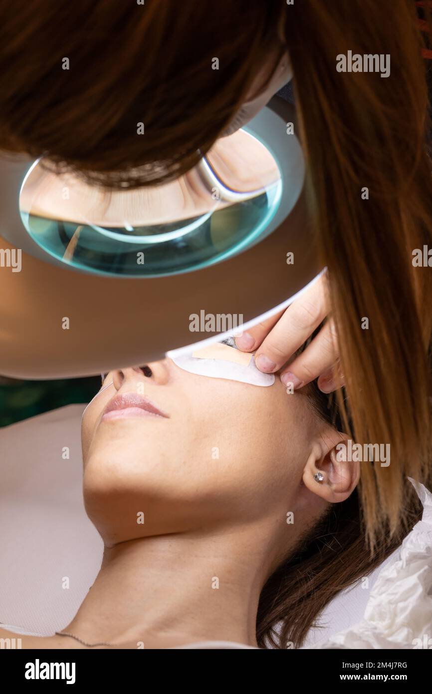 Junge Frau auf dem Bett des Kosmetikers für eine Wimpern-Verdickung. Nahaufnahme des Gesichts unter der Lampe mit Lupe Stockfoto