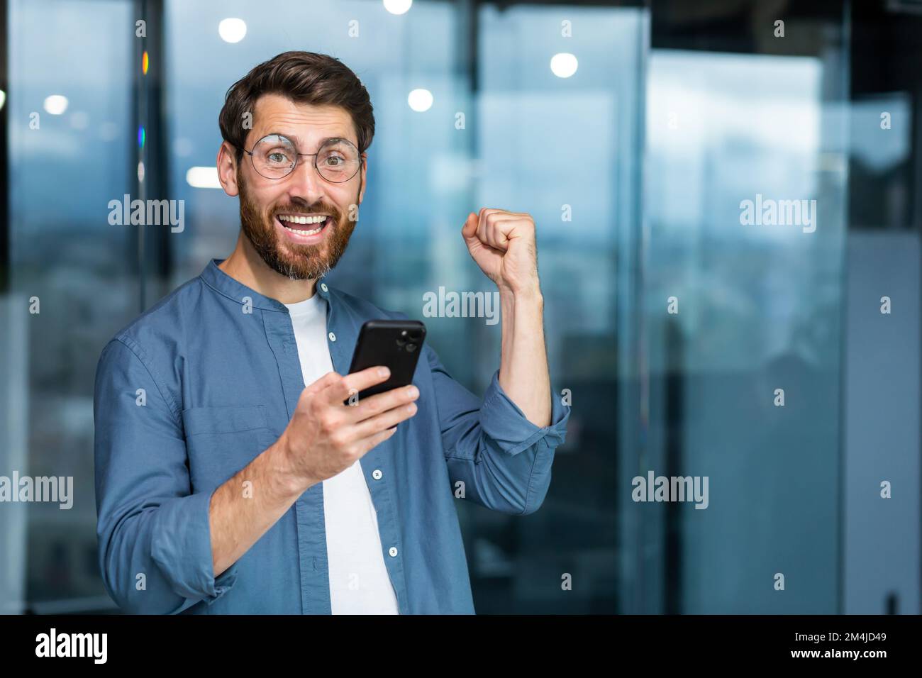 Porträt eines modernen Geschäftsmannes mitten im Büro, der Mann schaut mit einem Smartphone in die Kamera und feiert den Sieg, der Mann hält das Telefon in den Händen und hebt seine Hand in einer Geste des Triumphes. Stockfoto