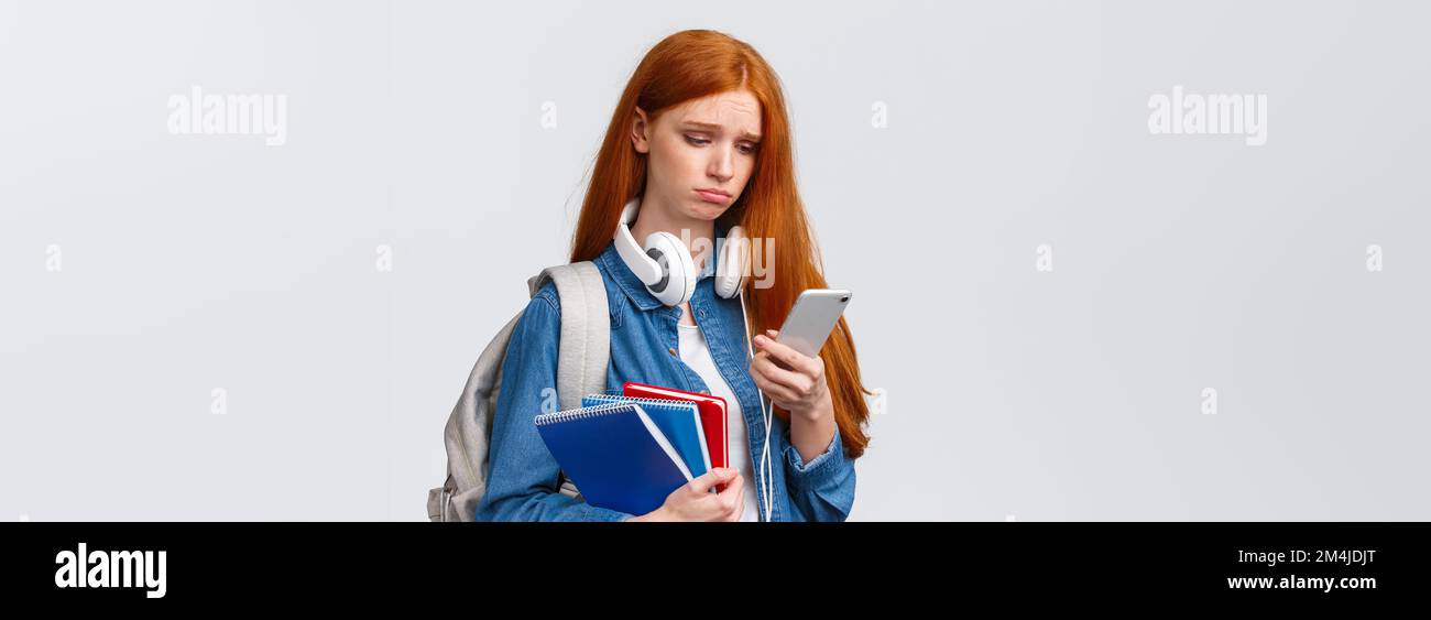 Albern trauriges und düsteres Pfadfindermädchen mit roten foxigen Haaren, schaurender Sichtung und Blick mit bedauern Smartphone-Bildschirm, Lesen störender Botschaft, Gefühl Stockfoto