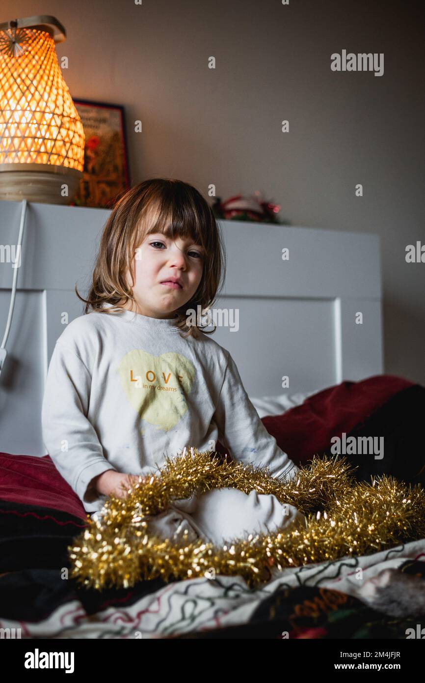 Aufgewühltes Mädchen in beigem Pyjama hat goldene weihnachtsdekoration auf dem Bett Stockfoto