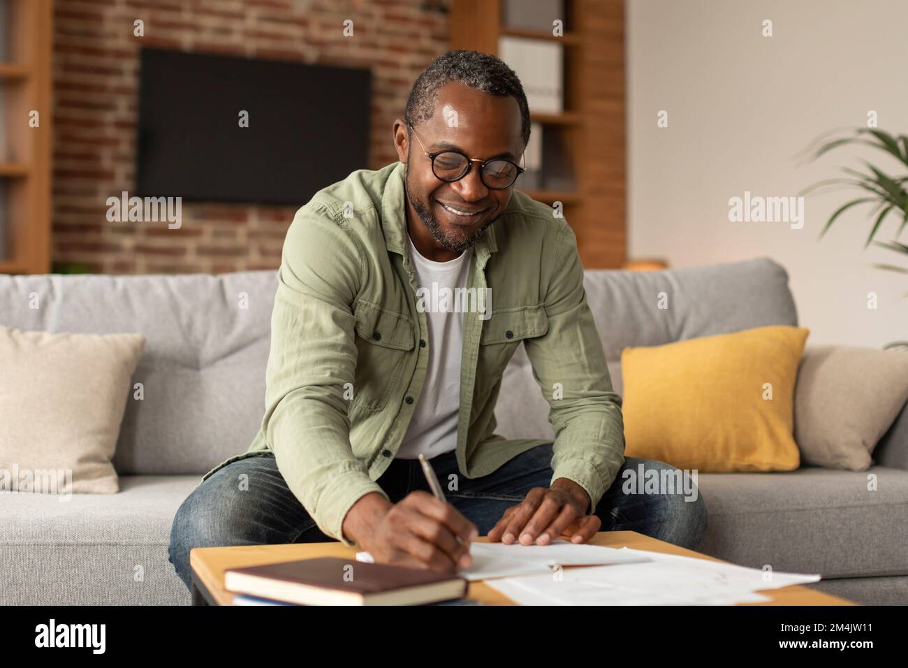 Ein fröhlicher schwarzer Geschäftsmann mit Brille macht Notizen, lernt und arbeitet im Wohnzimmer, im Kopierraum Stockfoto