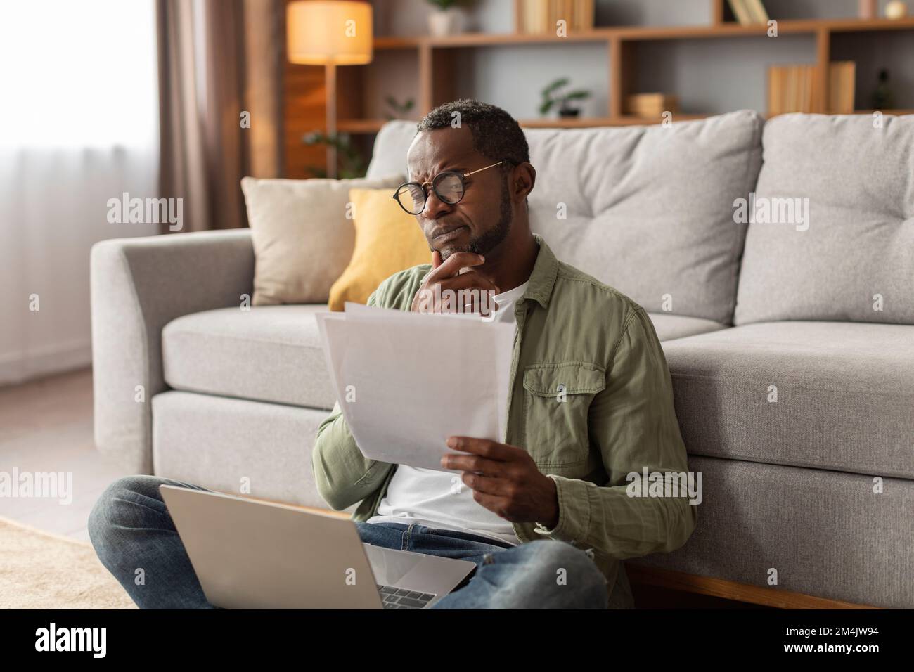 Der nachdenkliche Erwachsene Schwarze mit Brille benutzt Computer, arbeitet mit Dokumenten, sitzt auf dem Boden im Wohnzimmer Stockfoto