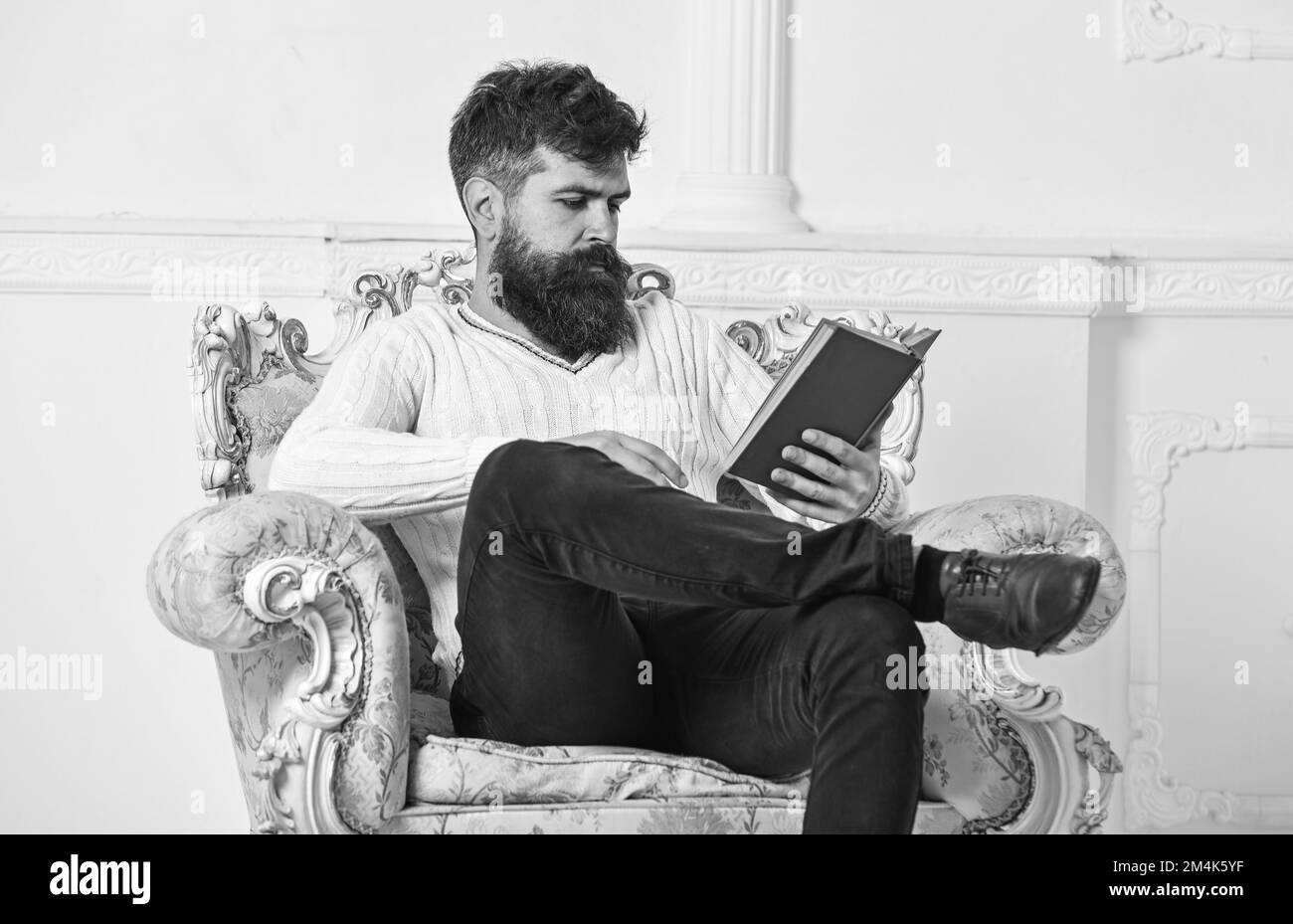Intelligentes Konzept. Wissenschaftler, Professor für Nachdenklichkeit, erforscht Literatur. Ein Mann mit Bart und Schnurrbart sitzt auf dem Sessel und liest Buch, weiße Wand Stockfoto