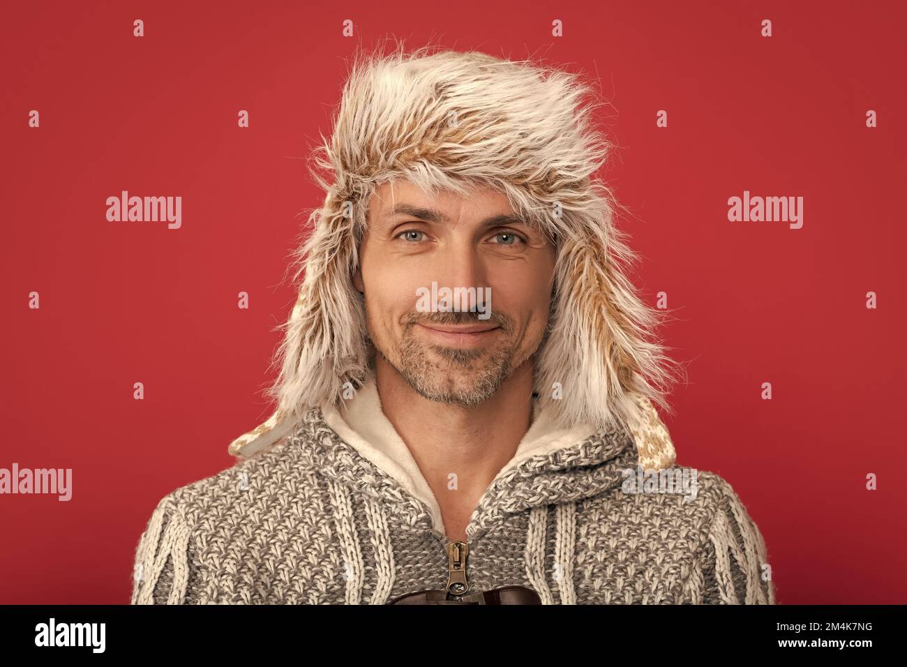 Lächelnder Grizzled Mann in Pullover und Ohrklappe auf rotem Hintergrund, Porträt Stockfoto