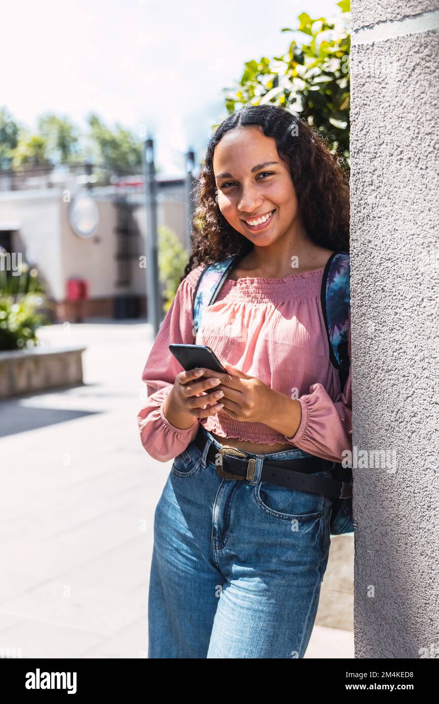 Lateinische Studentin, die lächelt und ihr Handy benutzt, um außerhalb des Campus in der Freizeit zu sitzen Stockfoto