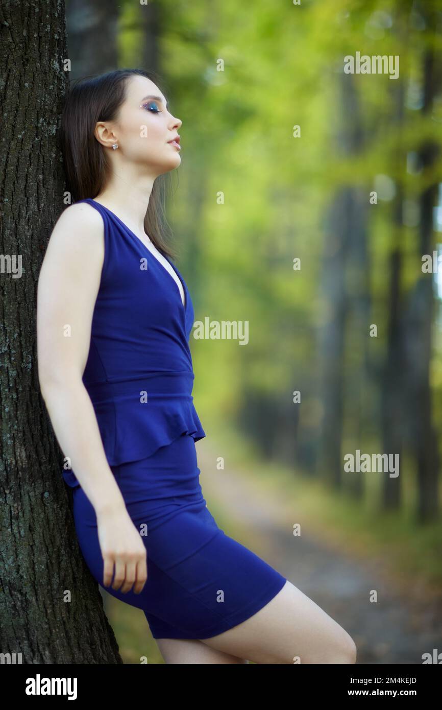 Eine Frau in blauem Kleid lehnt sich an einen Baumstamm in einer Herbstlindenbaumgasse. Pfad unter gelben Bäumen mit herbstlichen Blättern. Sonniger Herbsttag. Stockfoto