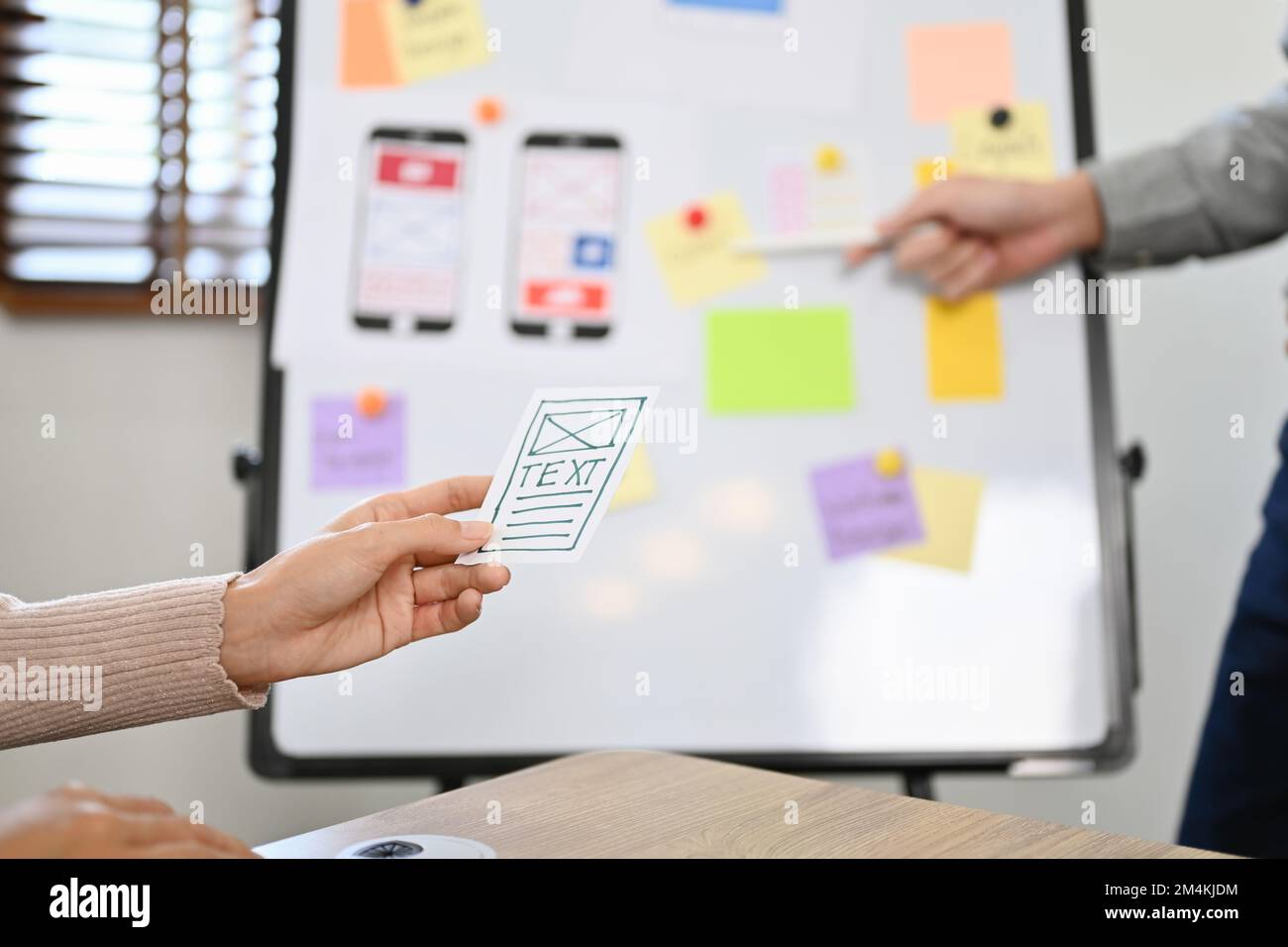 Eine weibliche Hand, die ein Designpapier für mobile Anwendungsschnittstellen über den unscharfen Hintergrund des Meetings hält. Zugeschnittenes Bild und Nahaufnahme Stockfoto