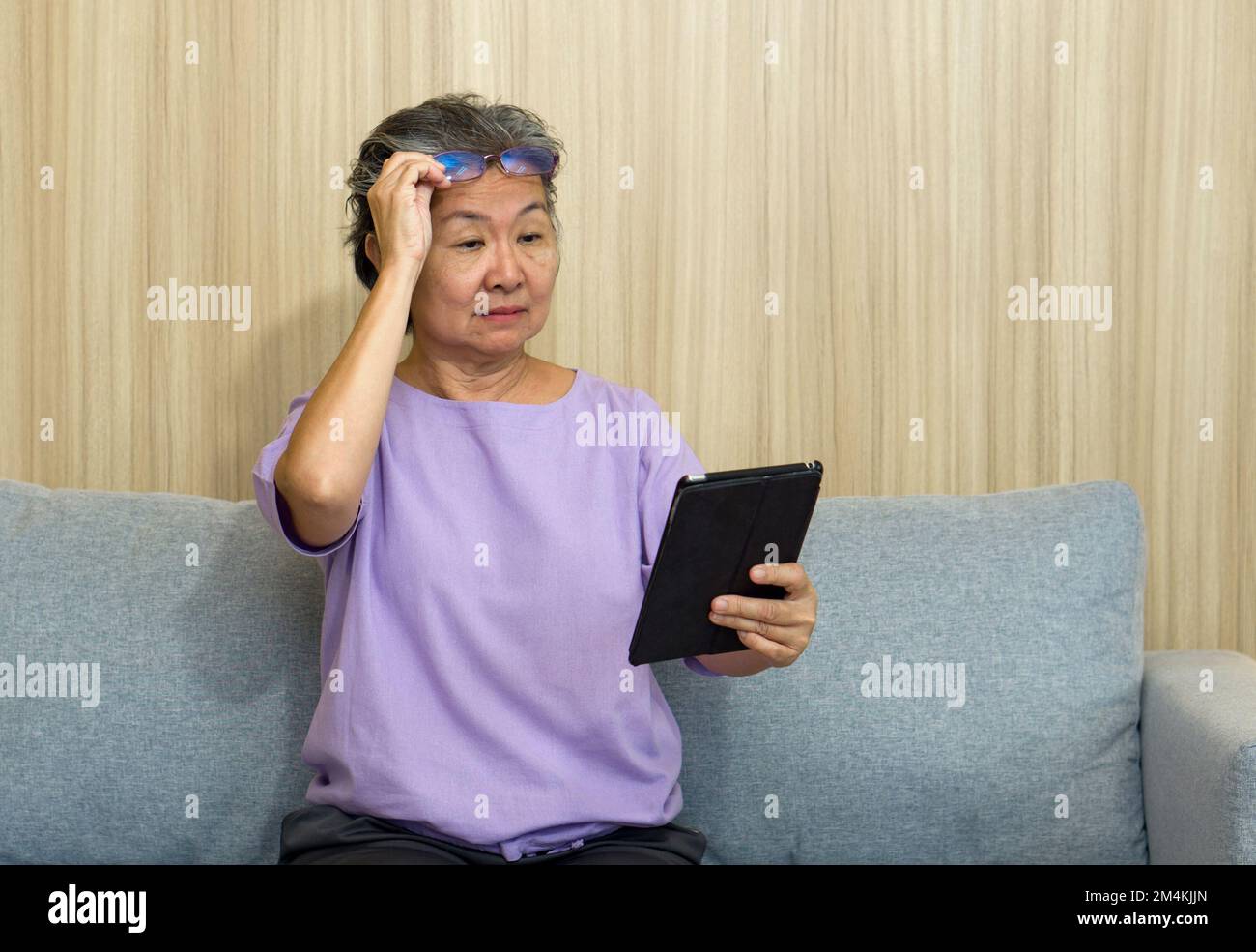 Ältere asiatische Rentner mit Brille sitzen auf dem Sofa und beobachten den Tablet-Computer. Mit Presbyopie-Syndrom kann sie sich nicht auf nahe gelegene Objekte konzentrieren. Stockfoto