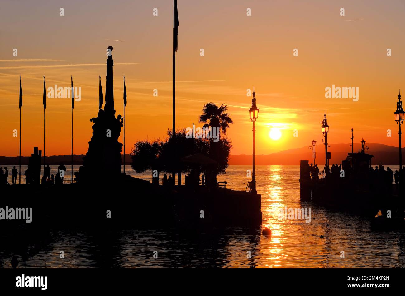 Lazise: Silhouette des Eingangs zum Fischereihafen mit Menschen kurz vor Sonnenuntergang in Lazise am See (Lago) Garda, Verona, Veneto, Italien Stockfoto