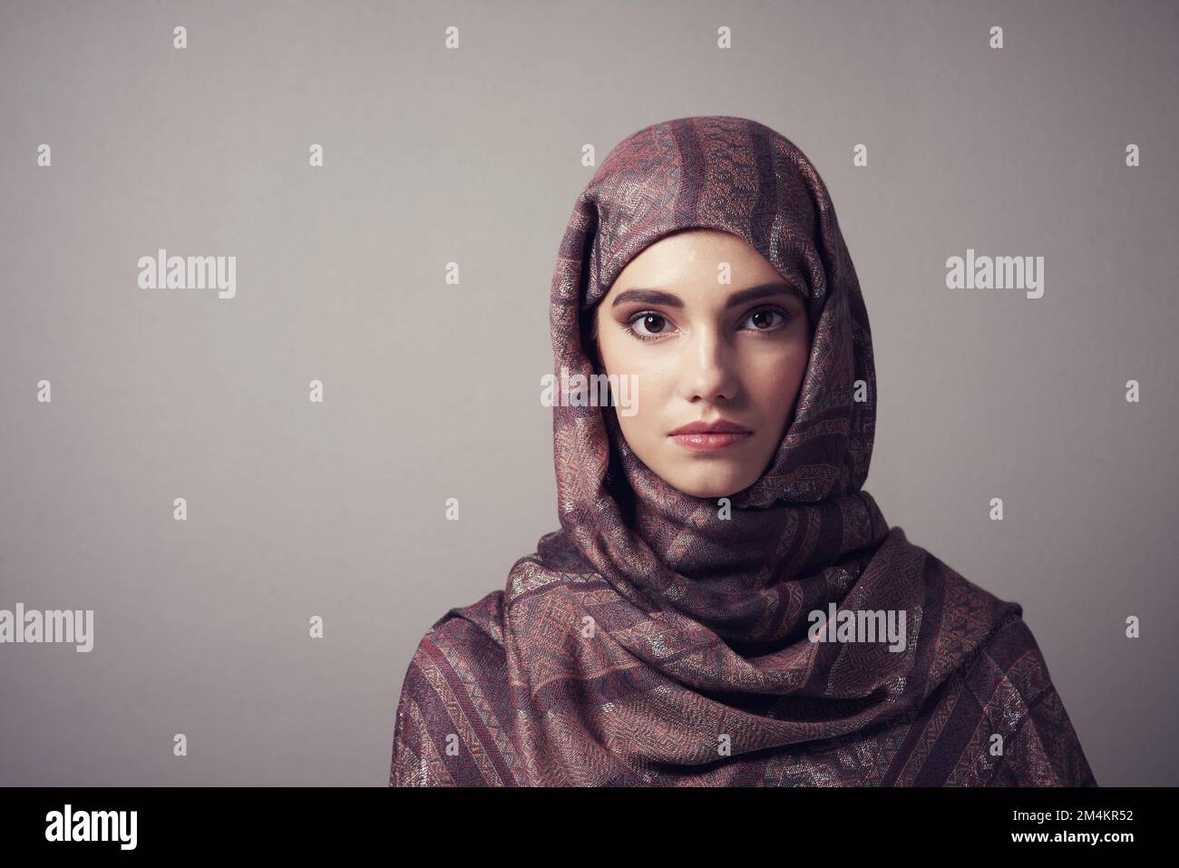 Erstaunlich selten. Studioporträt einer fröhlichen jungen Frau, die ein farbenfrohes Kopftuch trägt, während sie vor grauem Hintergrund posiert. Stockfoto