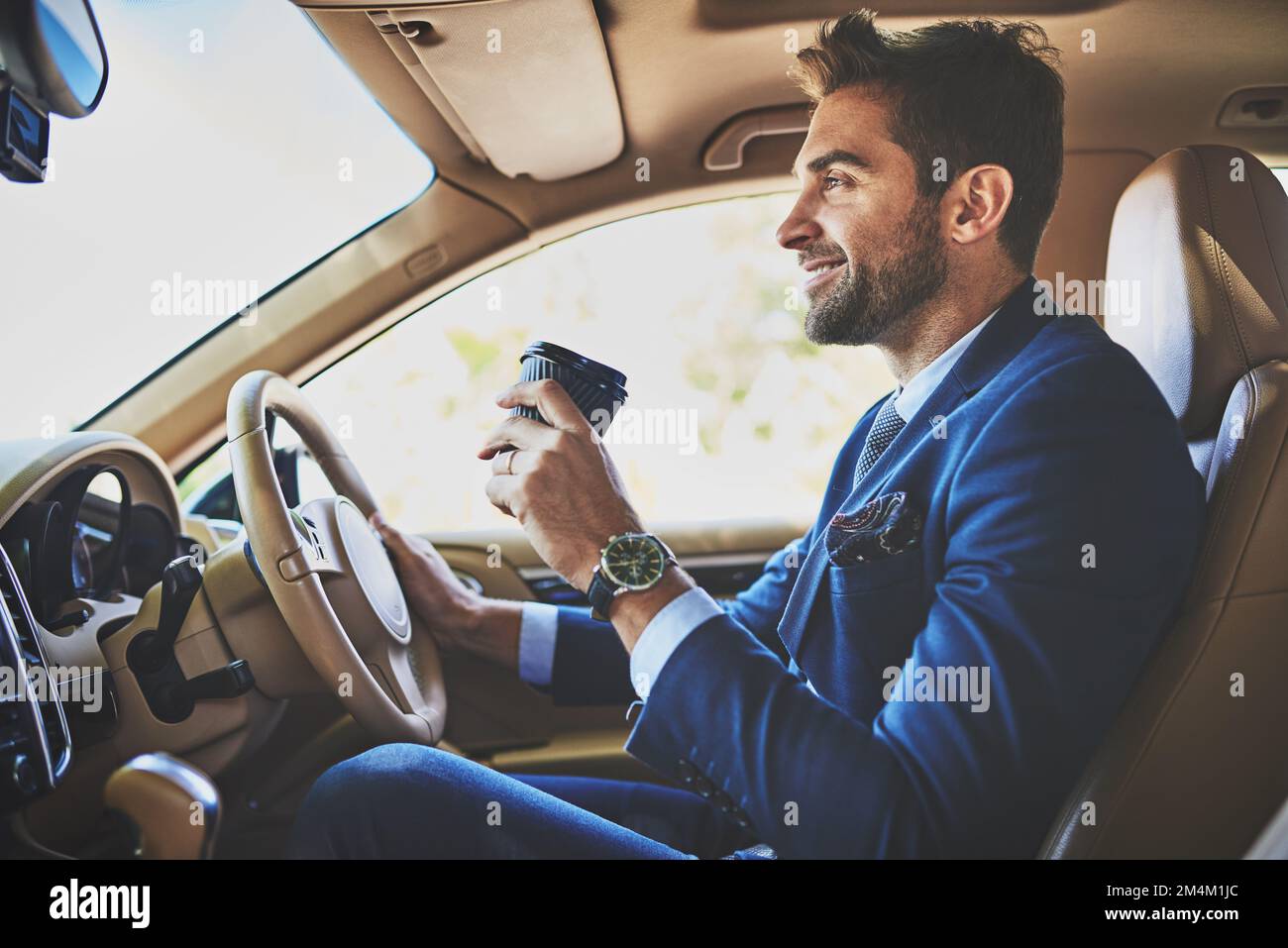 Nichts kann ihm die Stimmung verderben. Ein fröhlicher junger Geschäftsmann, der in seinem Auto zur Arbeit fährt, während er im Verkehr sitzt und Kaffee trinkt. Stockfoto