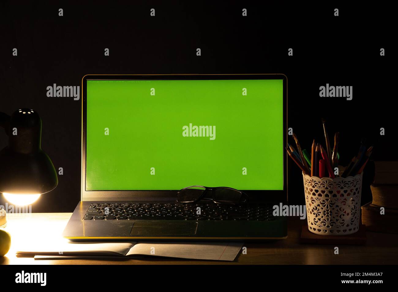 Laptop mit grünem Bildschirm auf dem Tisch mit Büchern in der Nähe der mitgelieferten hellen Lampe im dunklen Schlafzimmer zu Hause Stockfoto