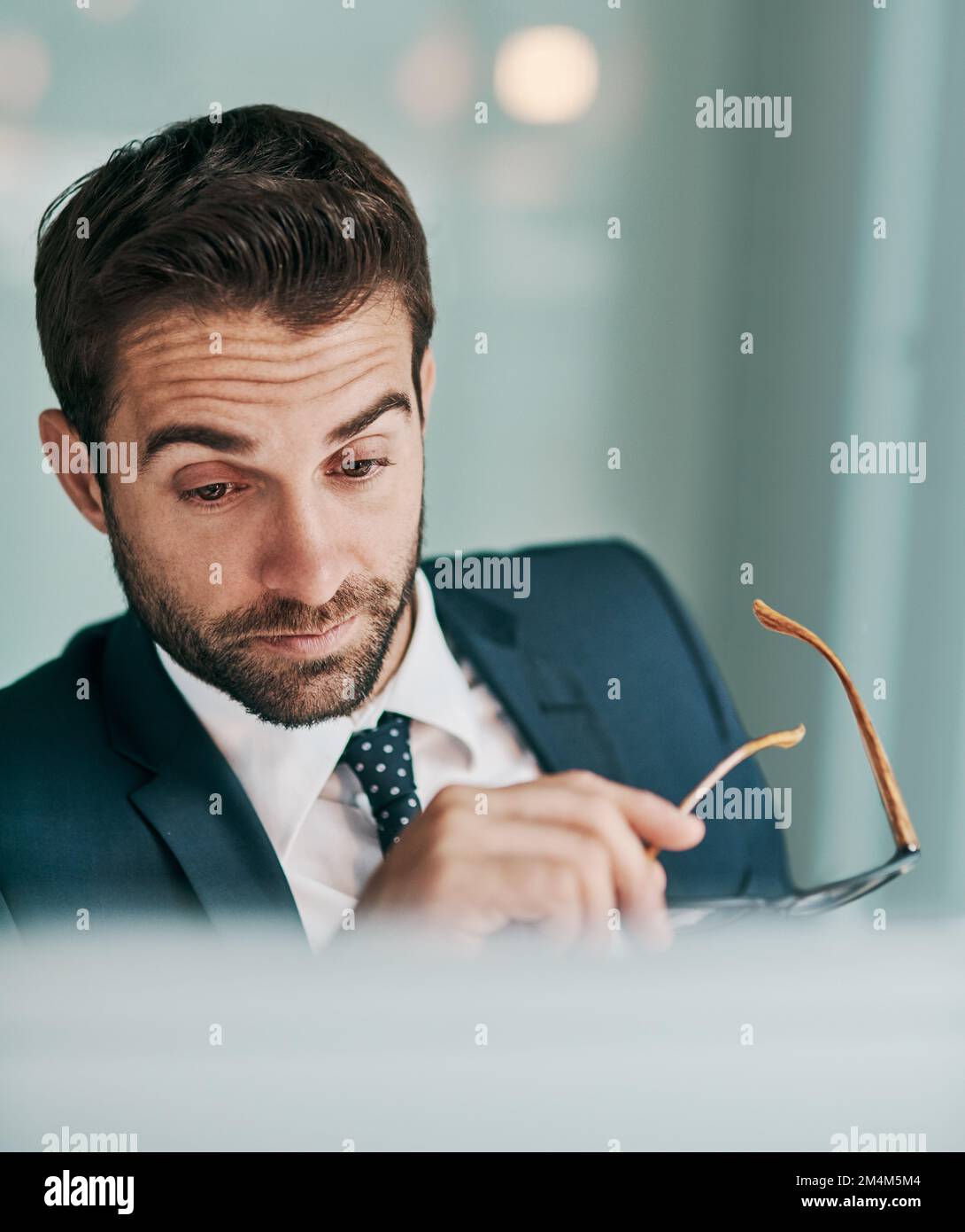 Die Arbeit stapelt sich immer mehr. Ein müder junger Geschäftsmann, der eine Brille hält, während er im Büro an einem Computer arbeitet. Stockfoto