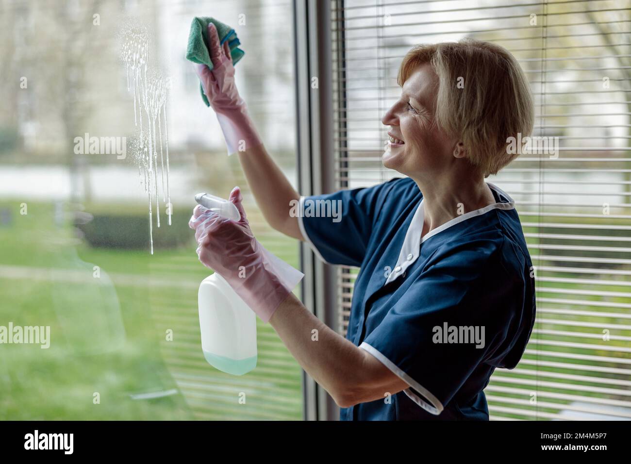 Lächelndes Hausmädchen in Handschuhen und einheitliches Reinigungsfenster mit Lappen und Reinigungsspray Stockfoto