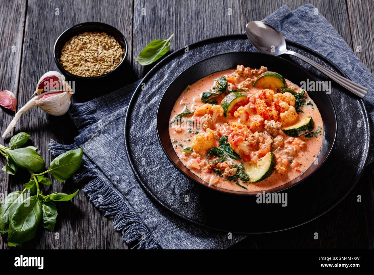 Cremige Tomatensuppe mit Hähnchen, Blumenkohl, Zucchini, Grünkohl, Basilikum und Gewürzen in schwarzer Schüssel auf dunklem Holztisch, horizontale Ansicht Stockfoto